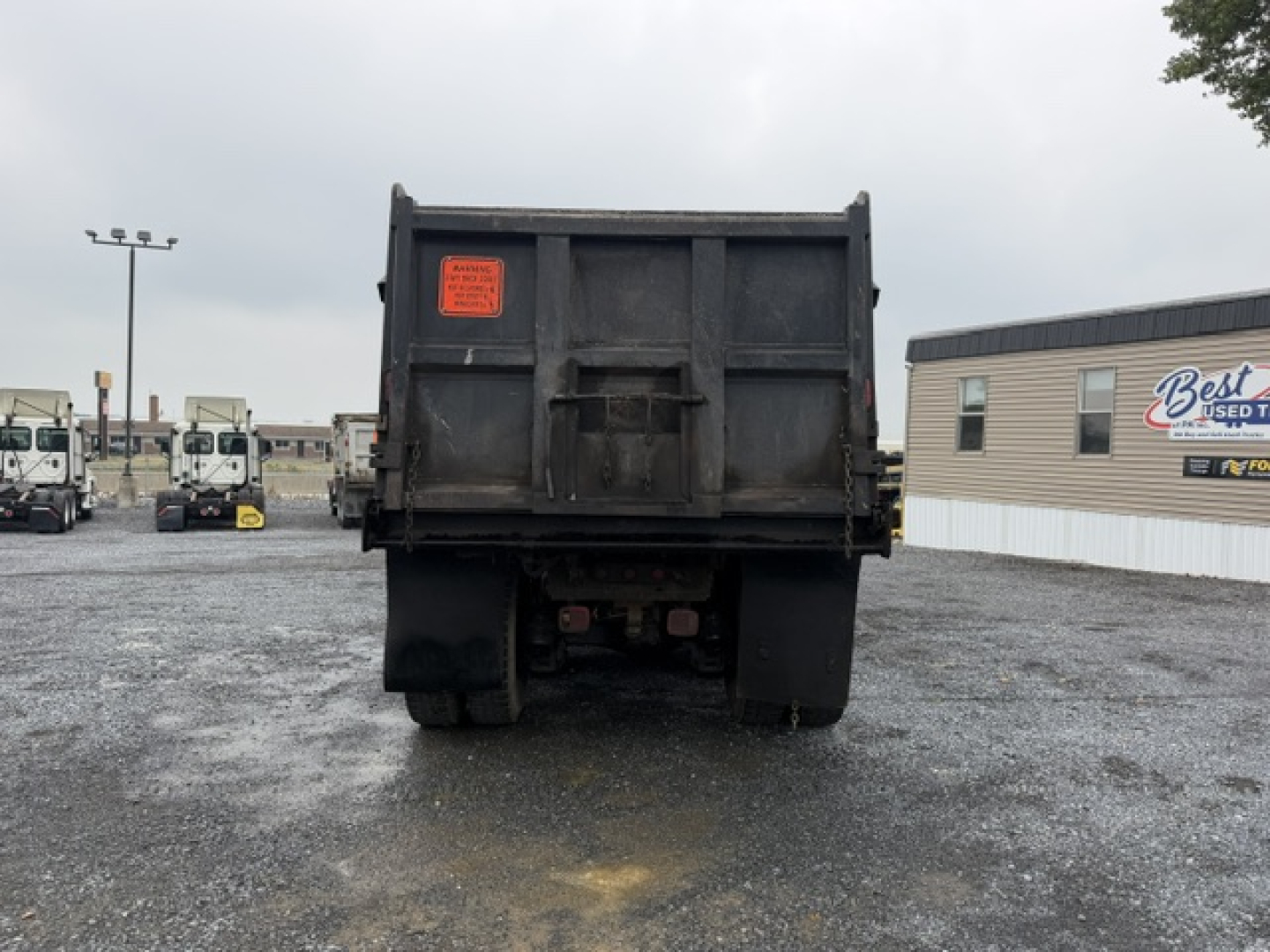 Rear view of a Mack CV713 dump truck with a black dump bed. An orange inspection label is on the left side of the bed. The truck is in a lot with other trucks and a building in the background.
