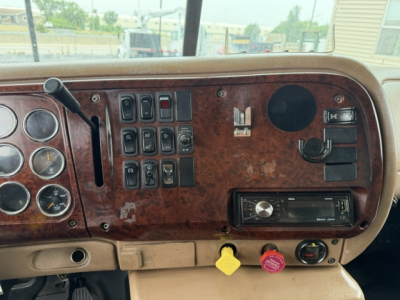 Dashboard interior of a Mack CV713 truck showing switches, gauges, and a radio with wood grain panel.