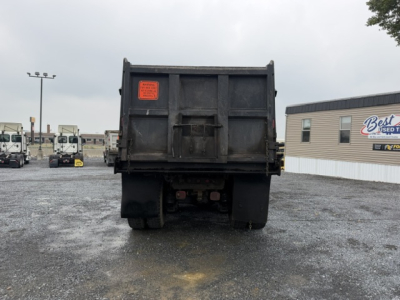 Rear view of a Mack CV713 dump truck with a black dump bed. An orange inspection label is on the left side of the bed. The truck is in a lot with other trucks and a building in the background.