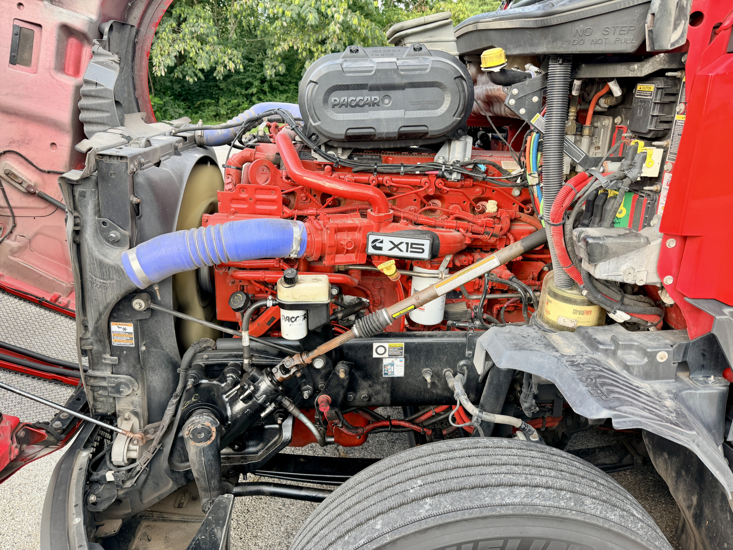 Kenworth T680 engine bay showing Cummins-? red engine with PACCAR badge, blue hose, various components and wiring in a truck cab interior area.