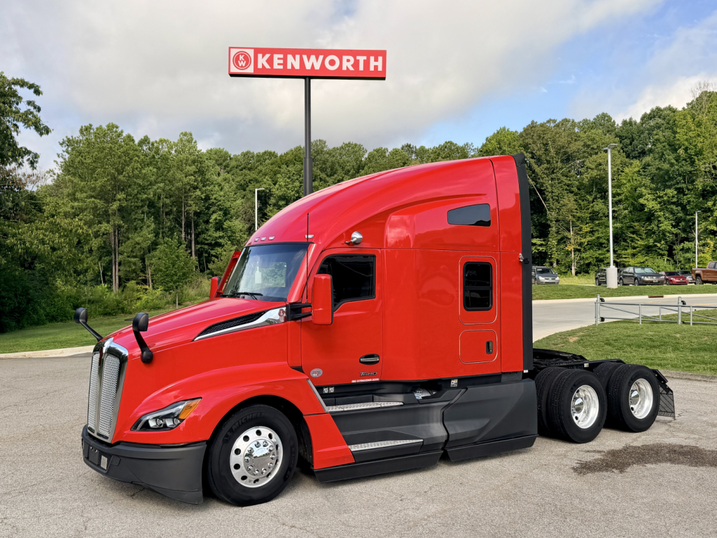 Kenworth T680 tractor in red, side view showing cab, sleeper, and 6x4 rear wheels, Kenworth sign in background.