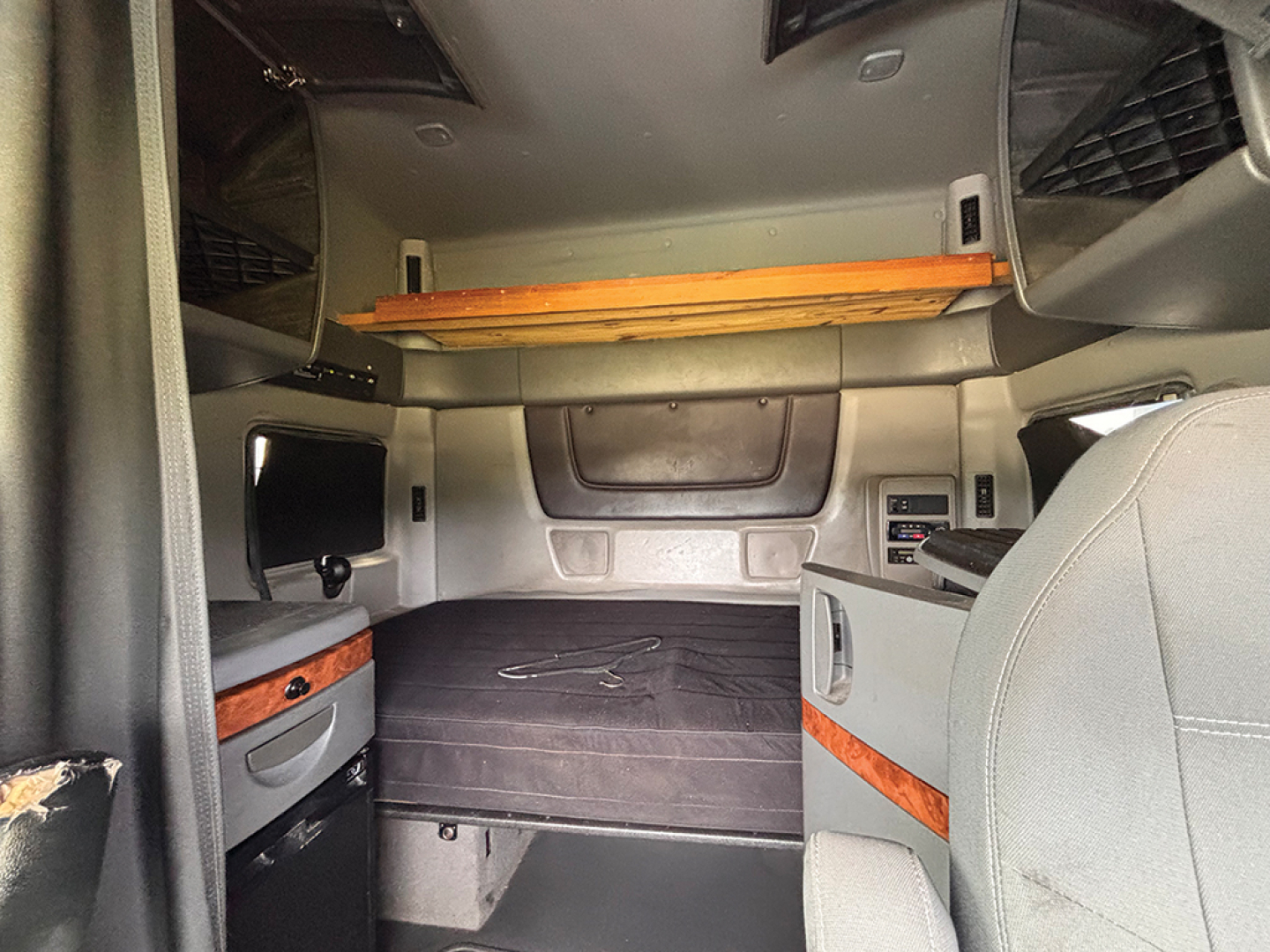 Interior of a truck sleeper cab with bed platform, overhead storage rack, and side compartments. Visible features include wood trim, grey upholstery, and control panels; no legible text appears in the scene.