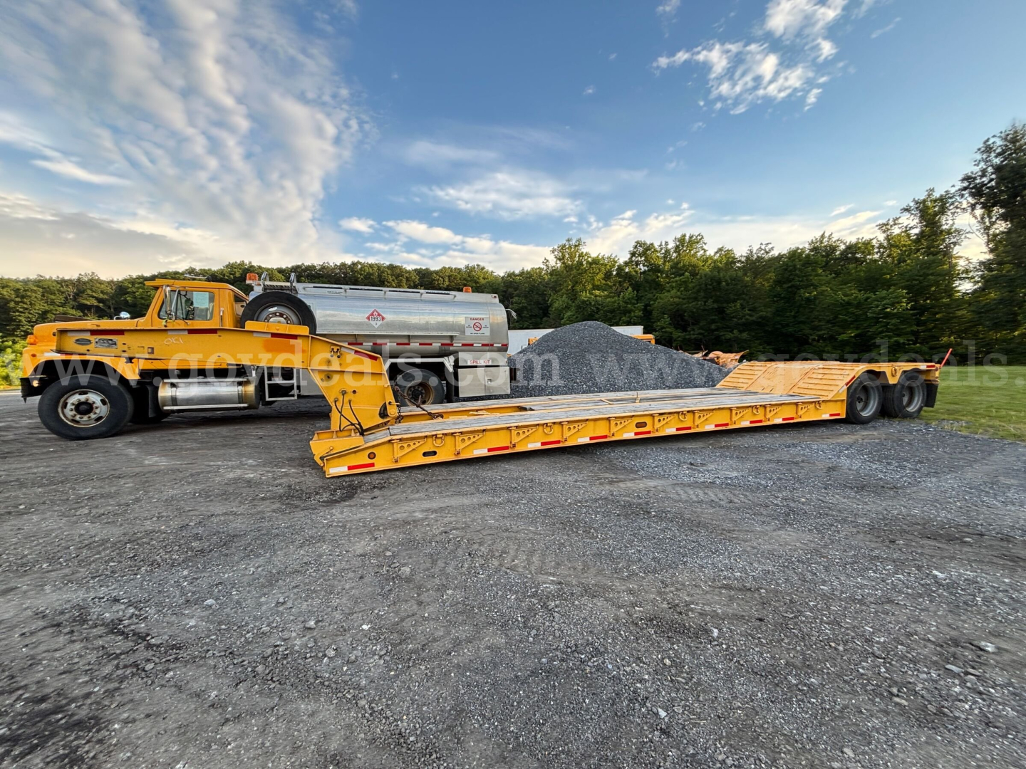 Lowboy trailer with a yellow gooseneck and lengthy flat deck loaded with gravel, attached to a yellow truck in a gravel lot.