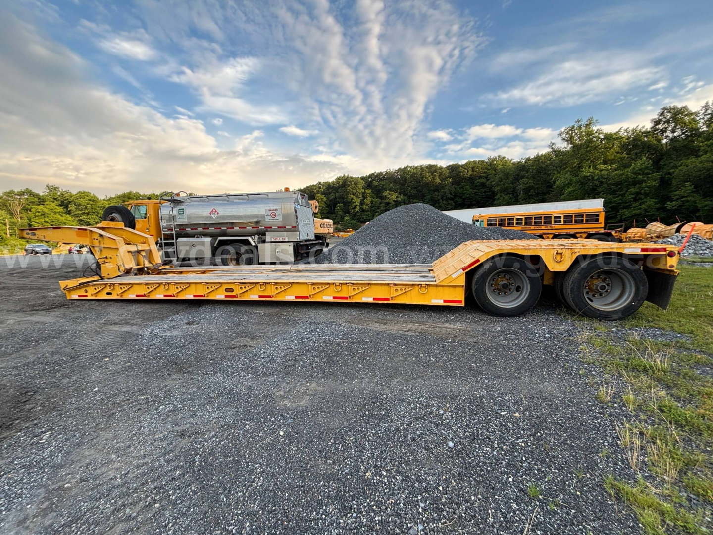 Rogers lowboy trailer in yard with gravel pile and pickup truck in background. Long flatbed with ramps, dual rear axles, yellow-orange color, and black tires.