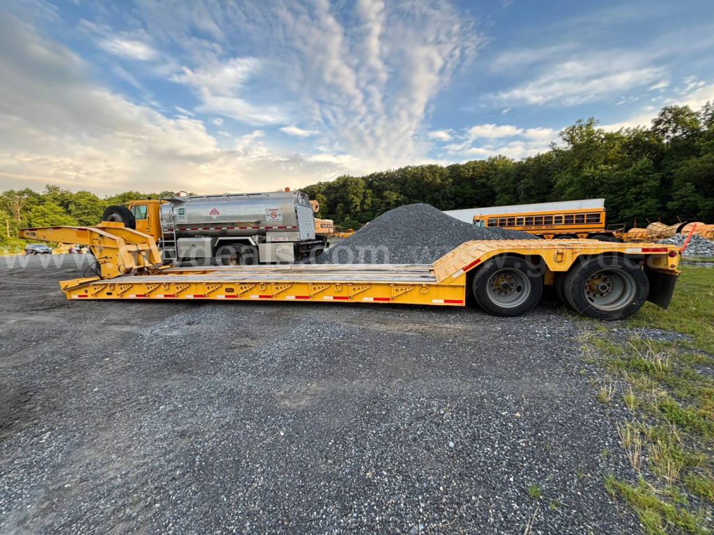 Rogers lowboy trailer in yard with gravel pile and pickup truck in background. Long flatbed with ramps, dual rear axles, yellow-orange color, and black tires.