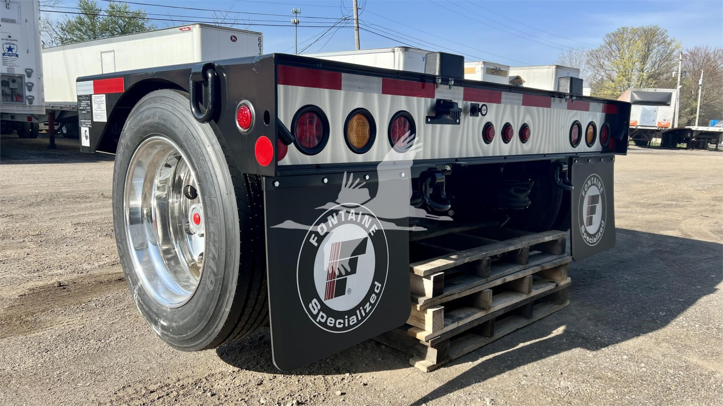 Fontaine Renegade lowboy trailer showing rear side with wheel, reflective stripes, circular logo mud flaps, and stacked wooden pallet undercarriage.