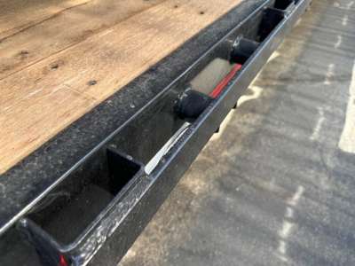 Drop deck trailer edge with wooden deck and black steel frame, showing stake pockets and mounting rails.