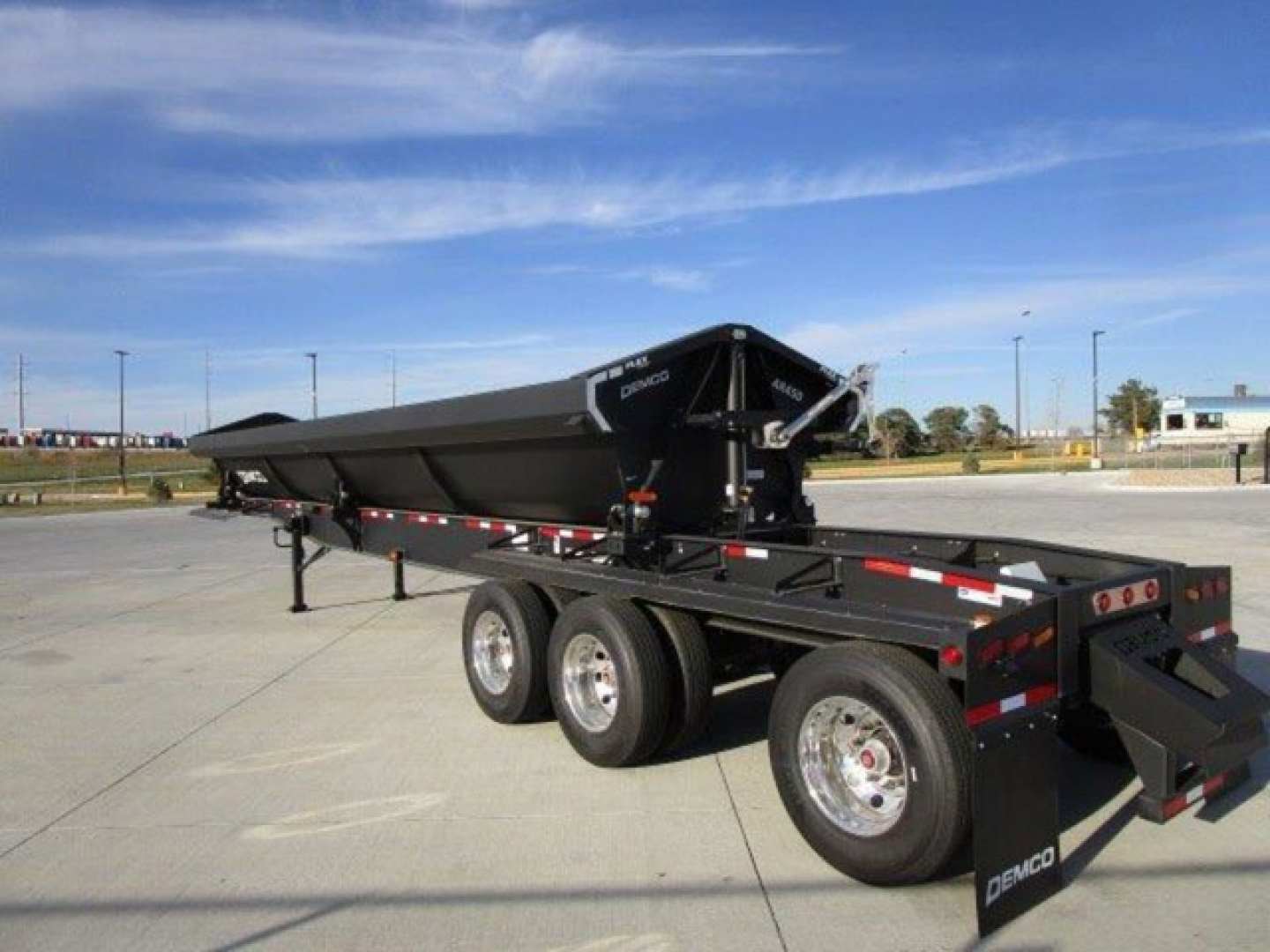 Demco dump trailer with powder-coated frame and dual-axle configuration, side rails, and rear drop-down tailgate. Brand name Demco visible on components; blue sky and dealership lot in background.