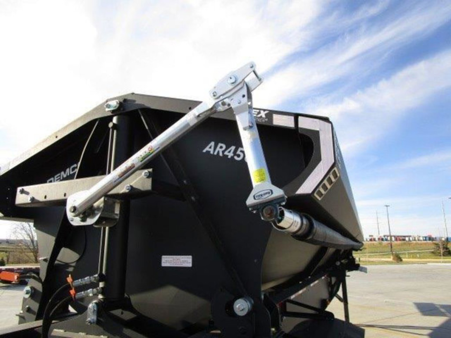 Dump trailer by Demco showing AR45 hitch linkage and hydraulic cylinder in black steel dump body with blue sky in background.