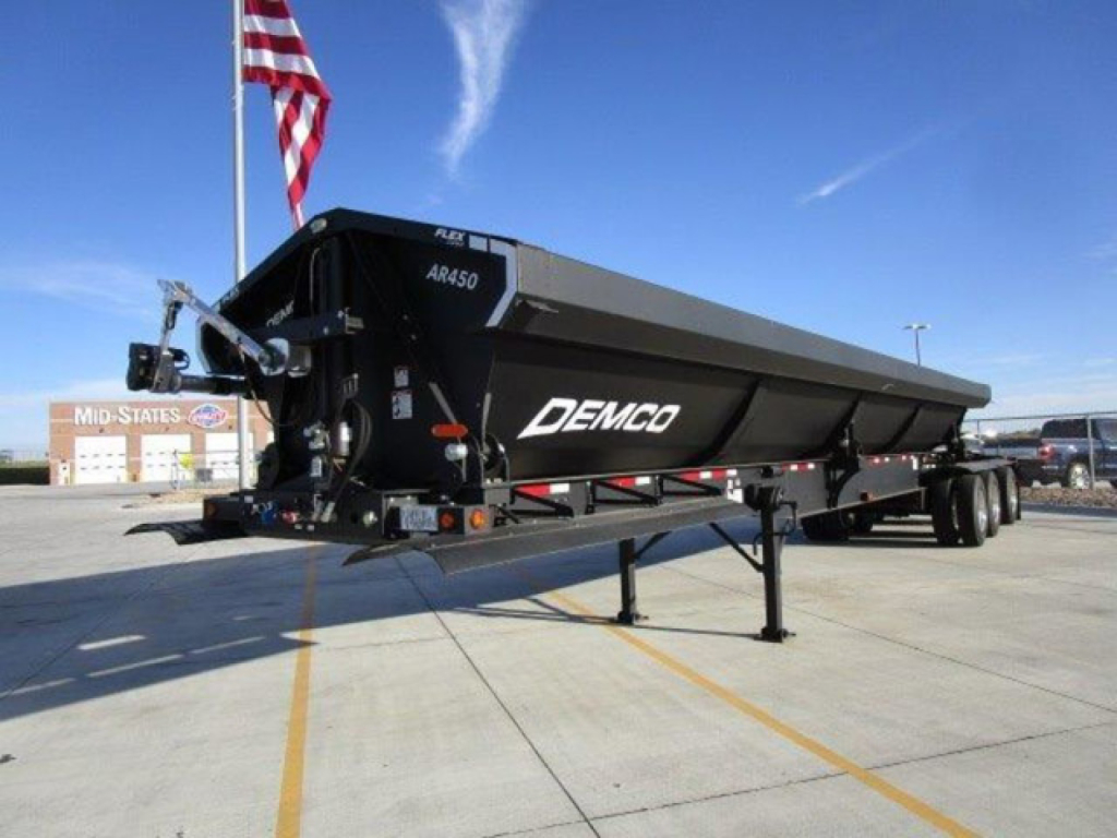Demco AR450 dump trailer in a parking lot viewed from front-left; black with white DEMCO logo, gooseneck hitch, landing gear, and dual rear axles; American flag in background.