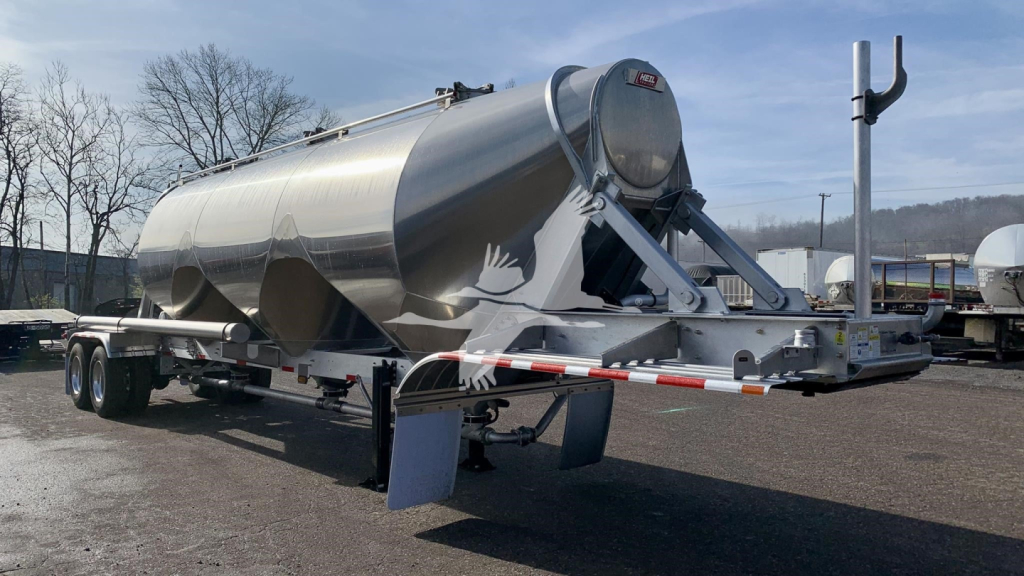Heil cementer trailer in a yard showing a polished aluminum cylindrical tank on a tri-axle chassis with a gooseneck hitch and landing gear.