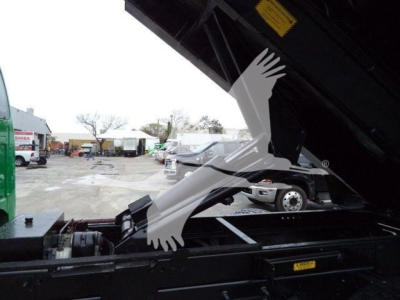 Truck bed with hydraulic dump tilt mechanism in foreground; partial view of cab and yard in background.