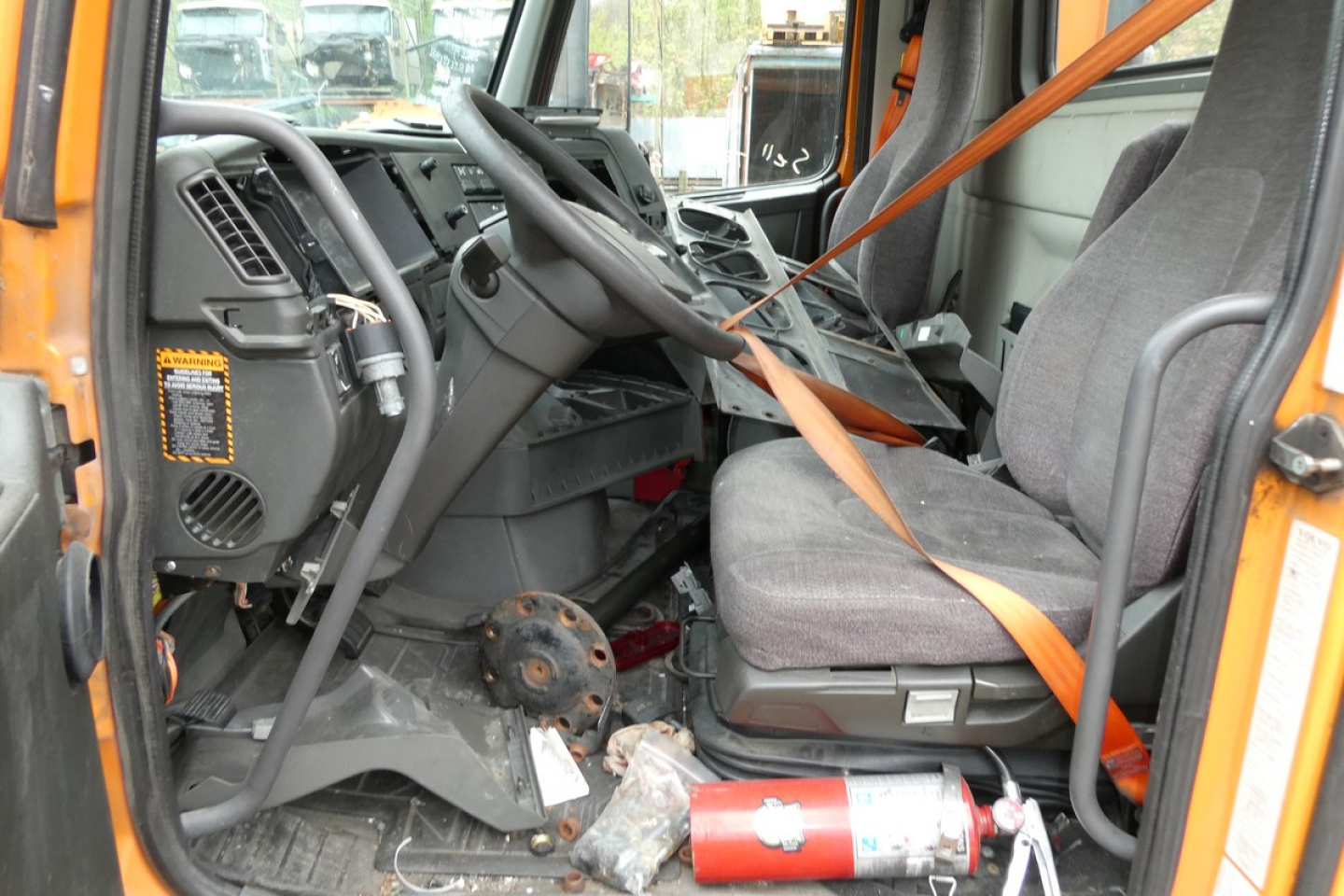 Volvo VNR64T300 truck cab interior showing gray driver's seat, steering wheel, dashboard, orange seat belt, and emergency fire extinguisher on the floor.