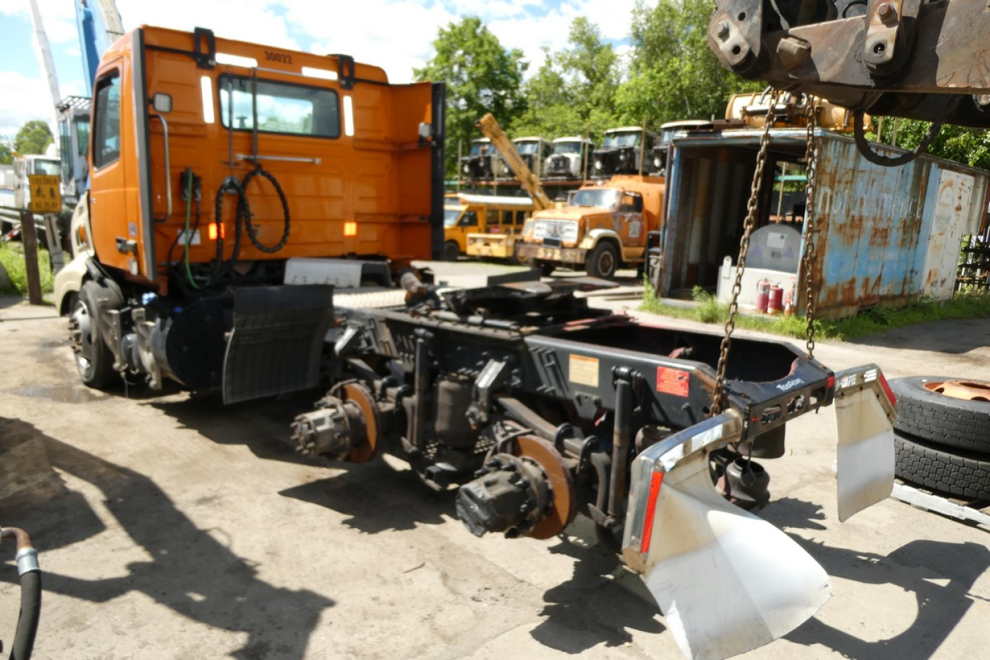 Volvo VNR64T300 chassis and cab seen in yard with detached rear drivetrain and mudflaps; orange sleeper cab in background.