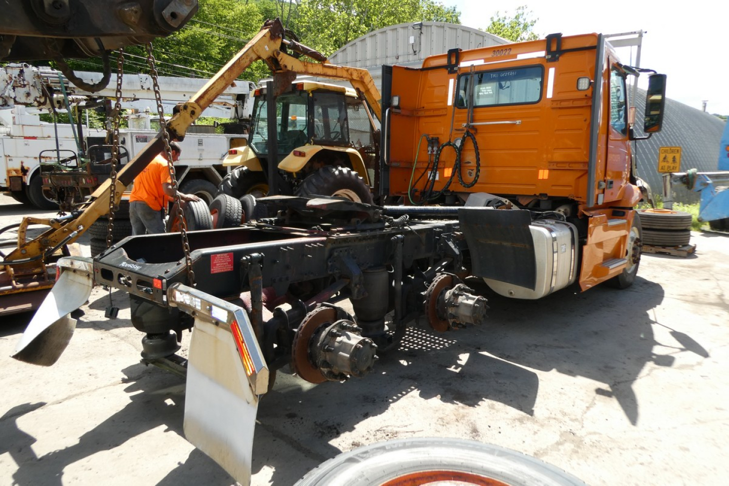 Vehicle chassis and cab from a Volvo VNR64T300. View shows the tractor with removed trailer connections, orange sleeper cab, chassis frame, axles, and hydraulic hoist in a repair yard.