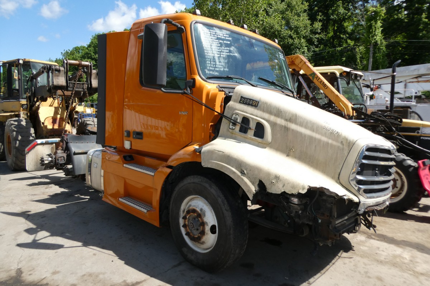 Volvo VNR64T300 cab and front chassis showing damaged right front fender and hood with visible windshield and side mirror, orange cab, junkyard background