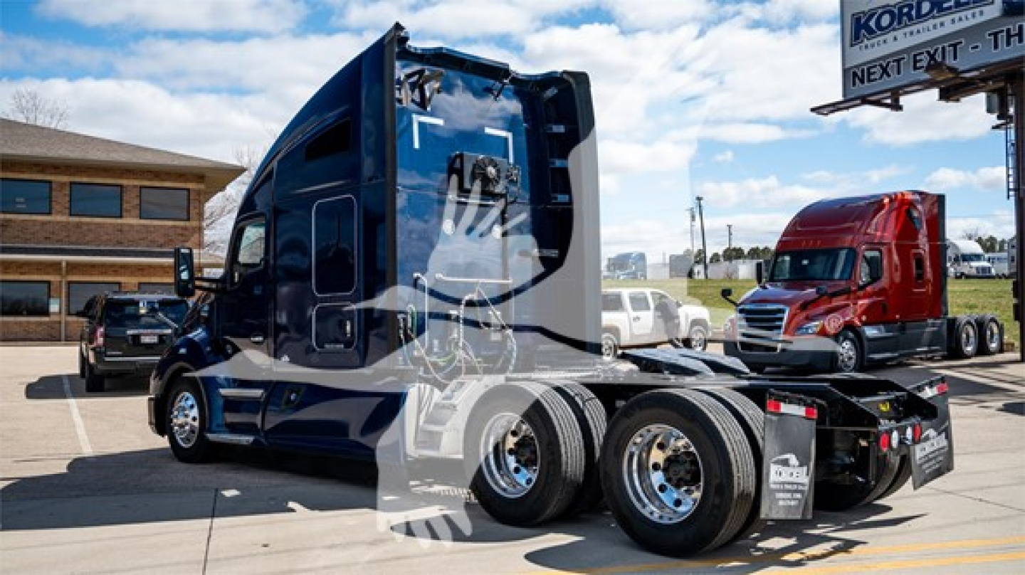 Kenworth T680 tractor-trailer in a lot showing side view, with multiple wheels and a sleeper cab, blue color, chrome rims, partial dealership lot in background.