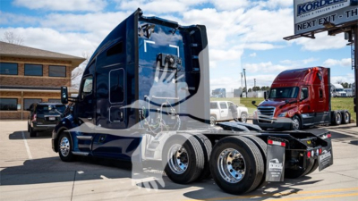 Kenworth T680 tractor-trailer in a lot showing side view, with multiple wheels and a sleeper cab, blue color, chrome rims, partial dealership lot in background.
