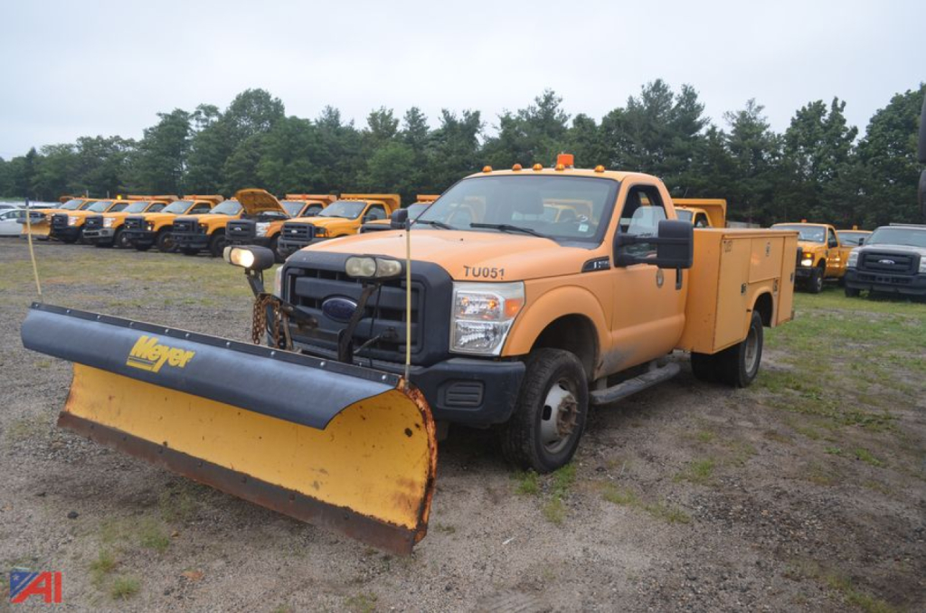 Ford F-350 Super Duty utility body pickup with snow plow attached, orange work truck in a lot of similar vehicles.
