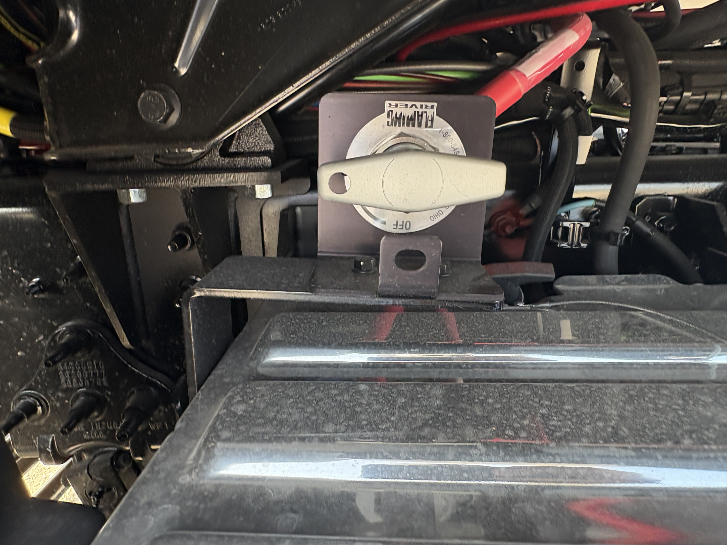 Truck engine bay area showing coolant reservoir with cap and surrounding hoses on a Mack GR64FT chassis, partial view of battery cables and metal components.