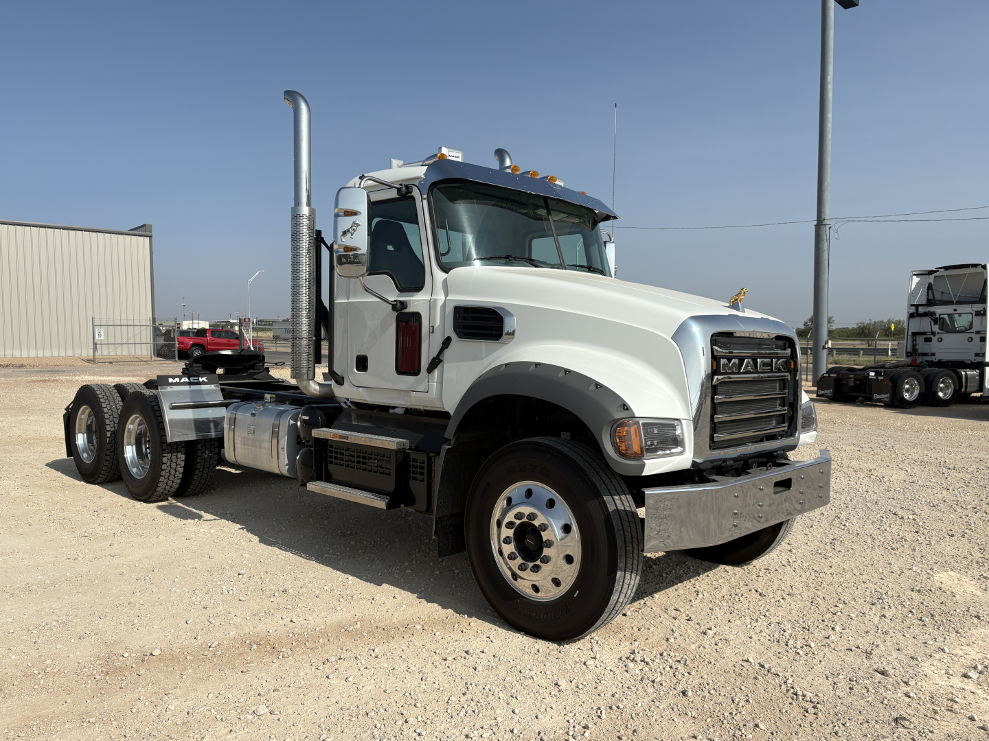 New white 2027 Mack GR64FT conventional daycab truck shown in a gravel lot, front three-quarter view with large chrome exhaust stack and grille, no trailer.