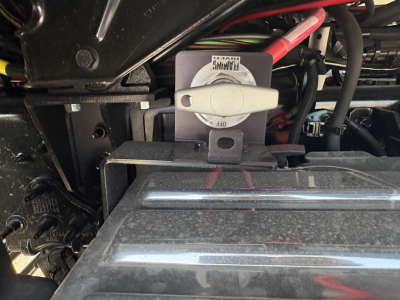 Truck engine bay area showing coolant reservoir with cap and surrounding hoses on a Mack GR64FT chassis, partial view of battery cables and metal components.