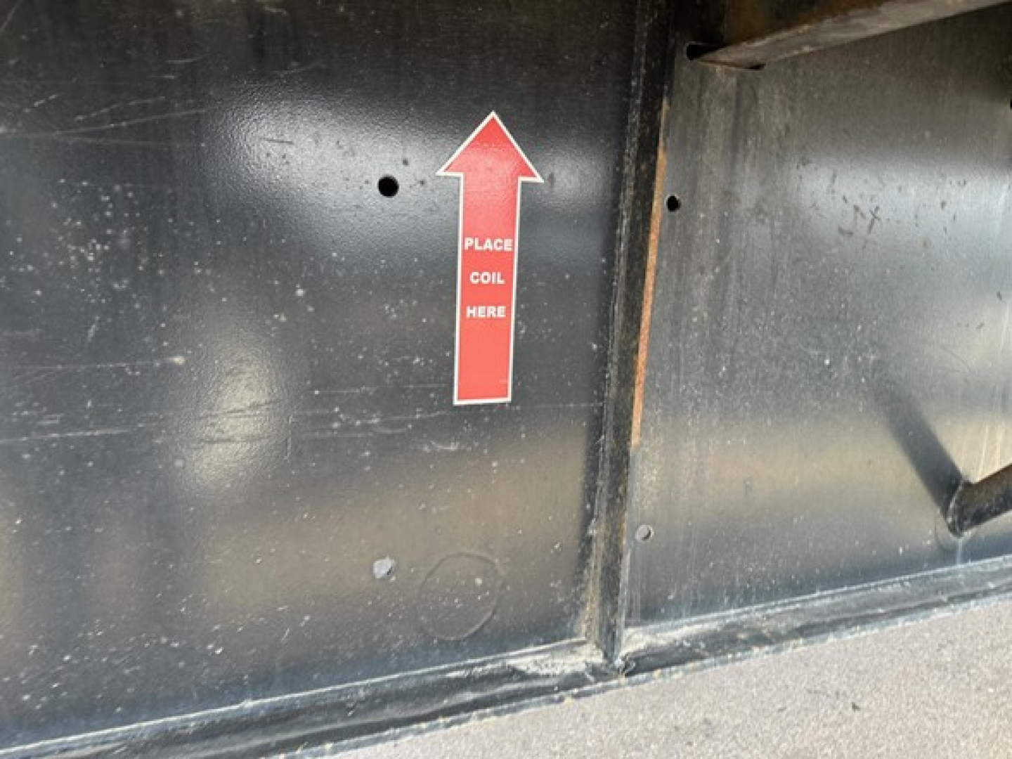 Flatbed trailer side showing a red up-arrow sticker with text PLACE COIL HERE on black metal surface.