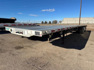 Flatbed trailer showing metal deck, dual rear axles, and landing gear with a wheel; visible side rails and safety reflectors along the edge.