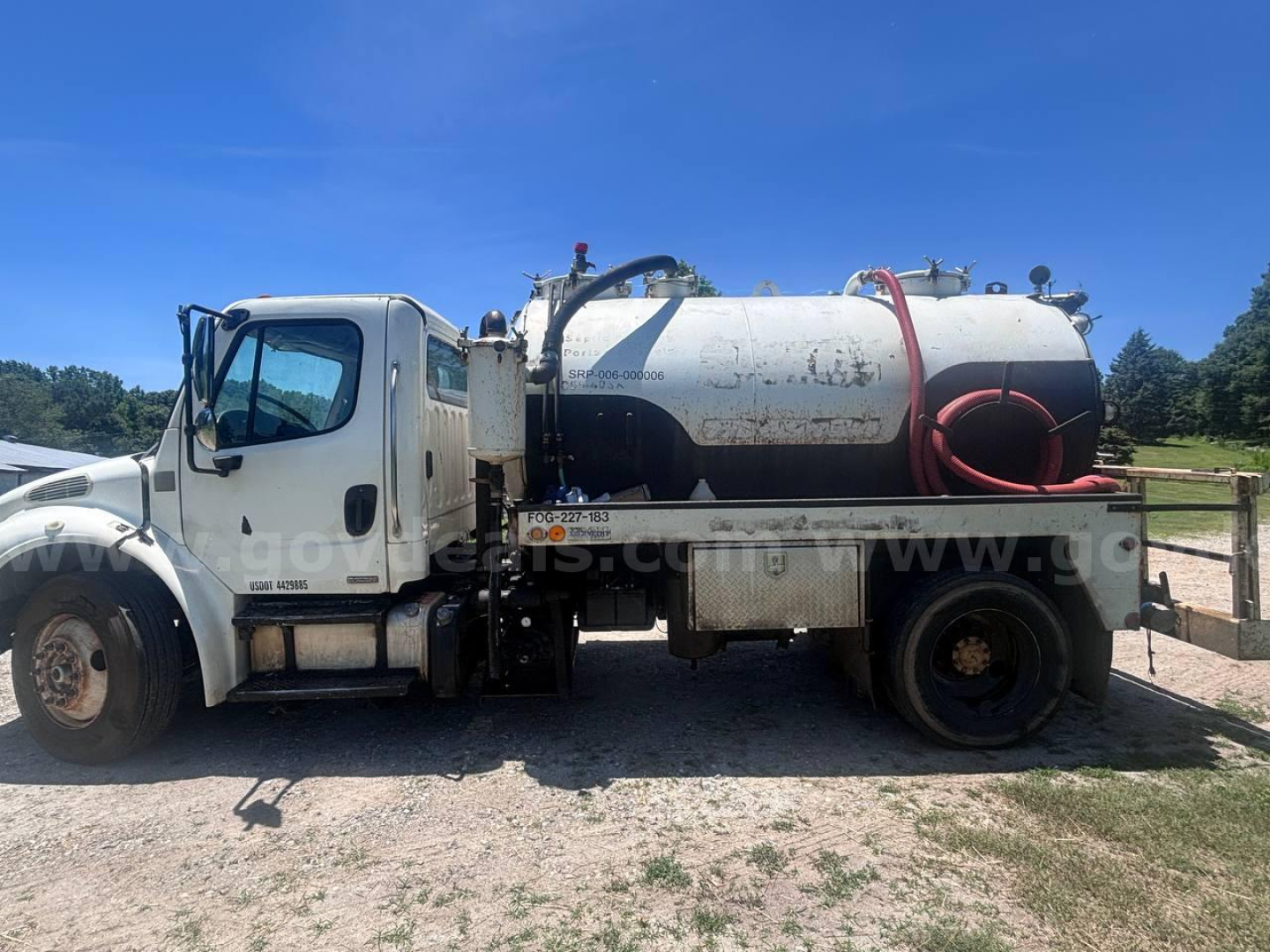 Truck-mounted vacuum tanker body on Freightliner M2 106, side view, white cab, cylindrical stainless tank with hoses and fittings, blue sky.