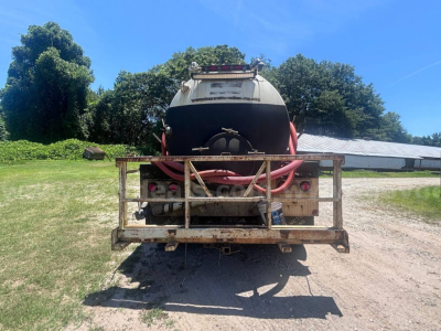 Rear view of a Freightliner M2 106 vacuum truck with a large cylindrical tank and red hoses, metal frame at tail, dirt road and green field in background.