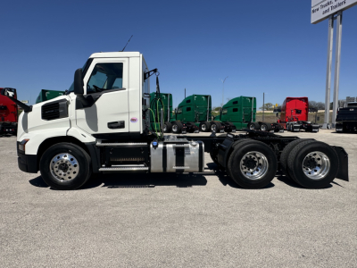 Truck chassis side view showing white Mack AN64T cab with dual rear axles and fuel tanks, highway trailer yard in background.