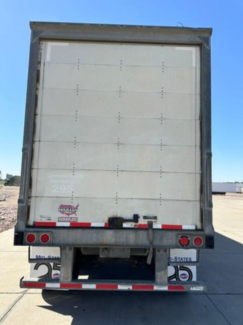 Rear view of a 2016 Wabash van trailer showing a large vertical roll-up door, rear bumper with lights and safety reflectors, and a hitch area. Visible text includes stock tag area signs and license plate-like stickers, but legible brand sti