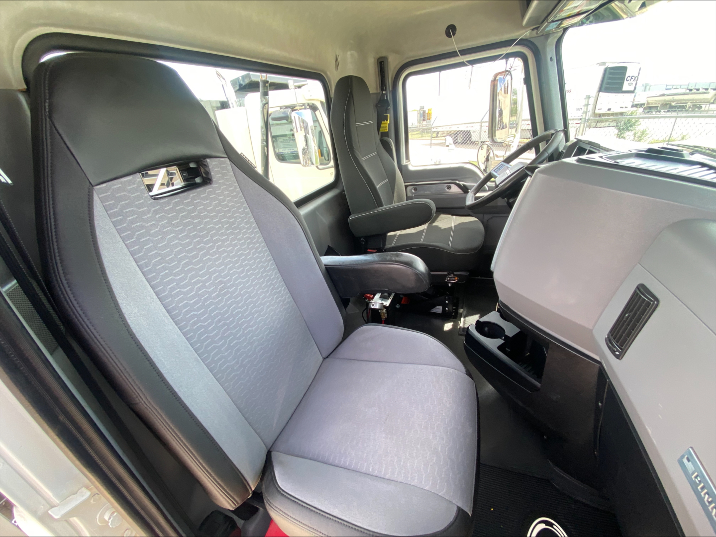 Truck cab interior of a 2021 Mack PI64T showing front bench seat, steering wheel, dashboard, and door panel with light gray upholstery.