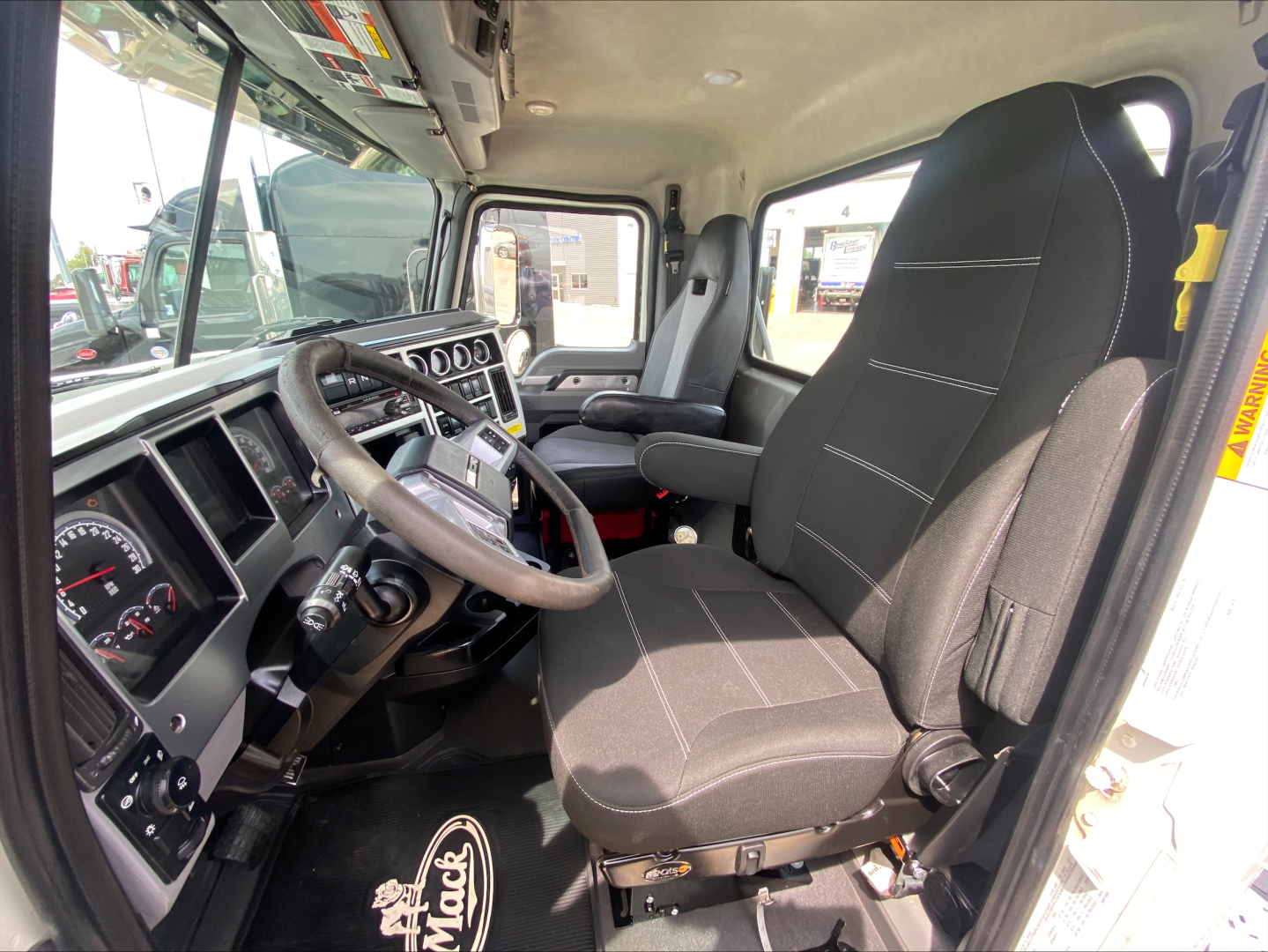 Truck cab interior of a 2021 Mack PI64T showing steering wheel, dashboard, driver seat, and passenger seat with center console.