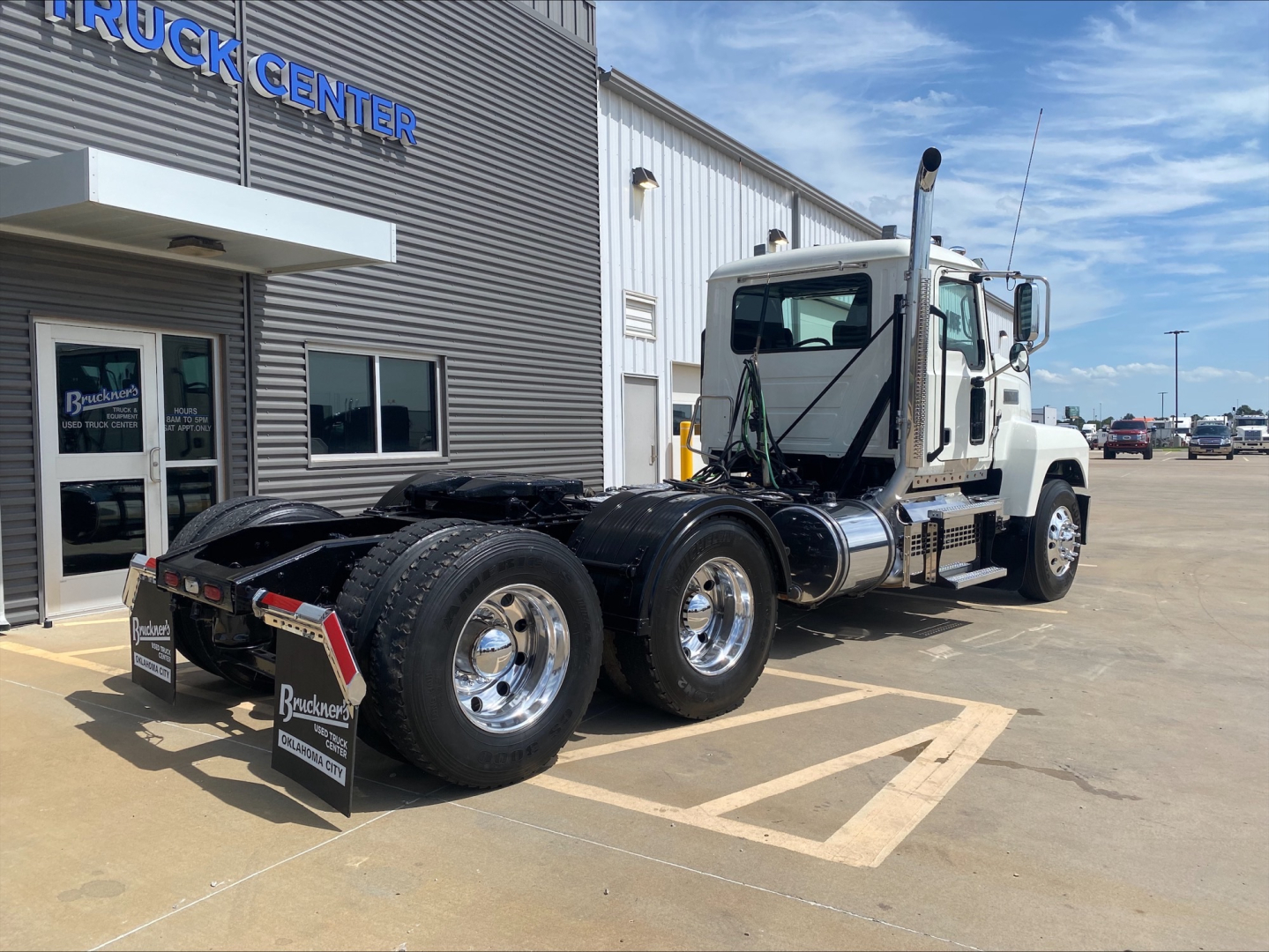 Truck tractor from a side-rear view showing a white Mack PI64T daycab with dual rear axles and black chassis at a dealership lot.