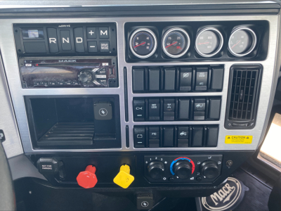 Dashboard panel of a 2021 Mack PI64T showing R N D + M shifter, gauges, climate controls, switches, and two colored knobs (red and yellow) with a Mack badge visible.