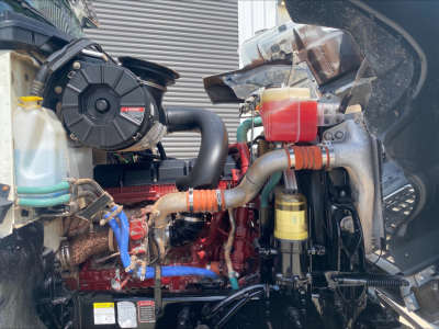 Mack PI64T engine bay showing coolant reservoir, turbocharger, hoses, and yellow oil filter with red engine block visible.