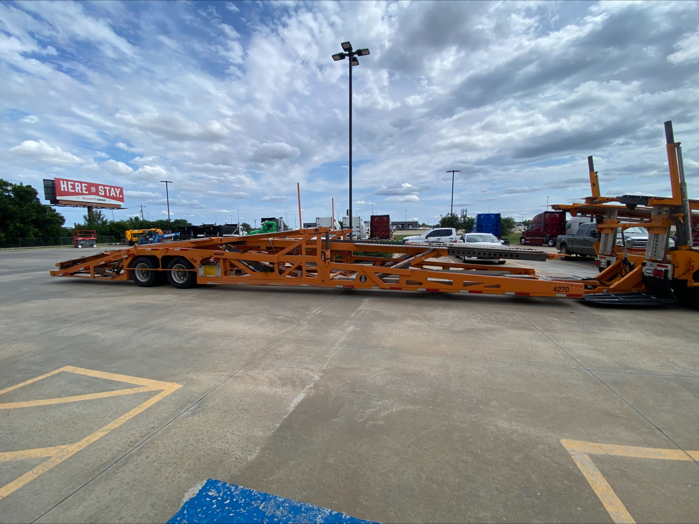 Volvo VAH auto carrier trailer in yard, long orange hydraulic tilt deck with dual rear axles and support frames, equipment and trucks in background.
