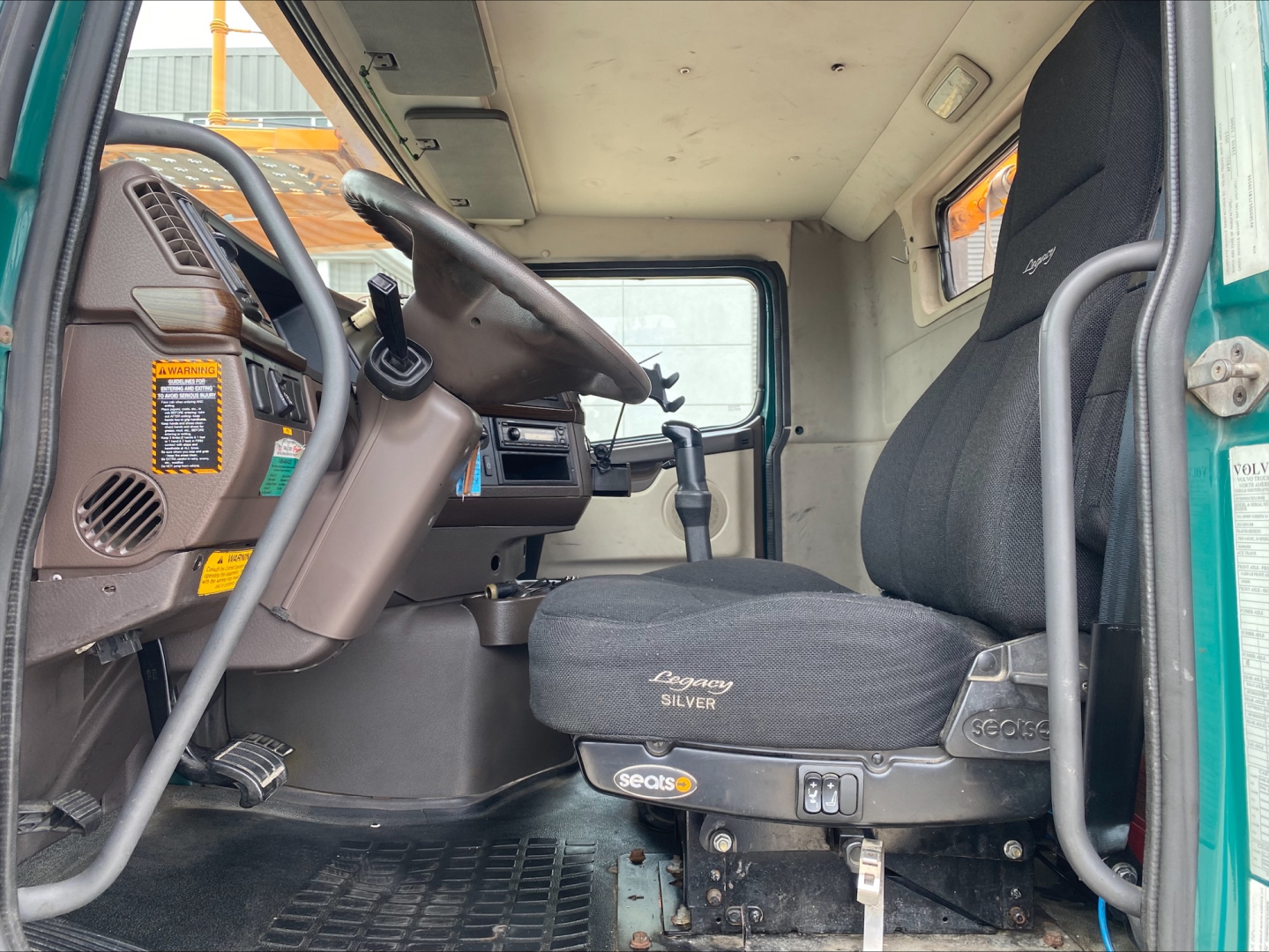 Truck cab interior of a 2014 Volvo VAH with steering wheel, grey driver seat, dash controls, and floor mat visible.