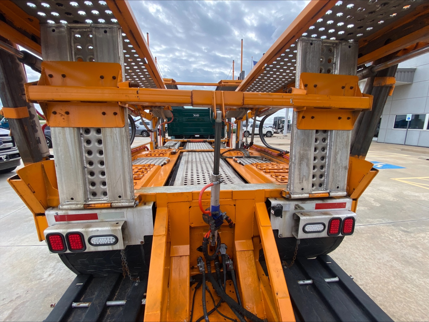 Industrial trailer with orange metal framework and dual rear lights visible; steel rails and hydraulic components centered; truck bed racks and ramp structure.