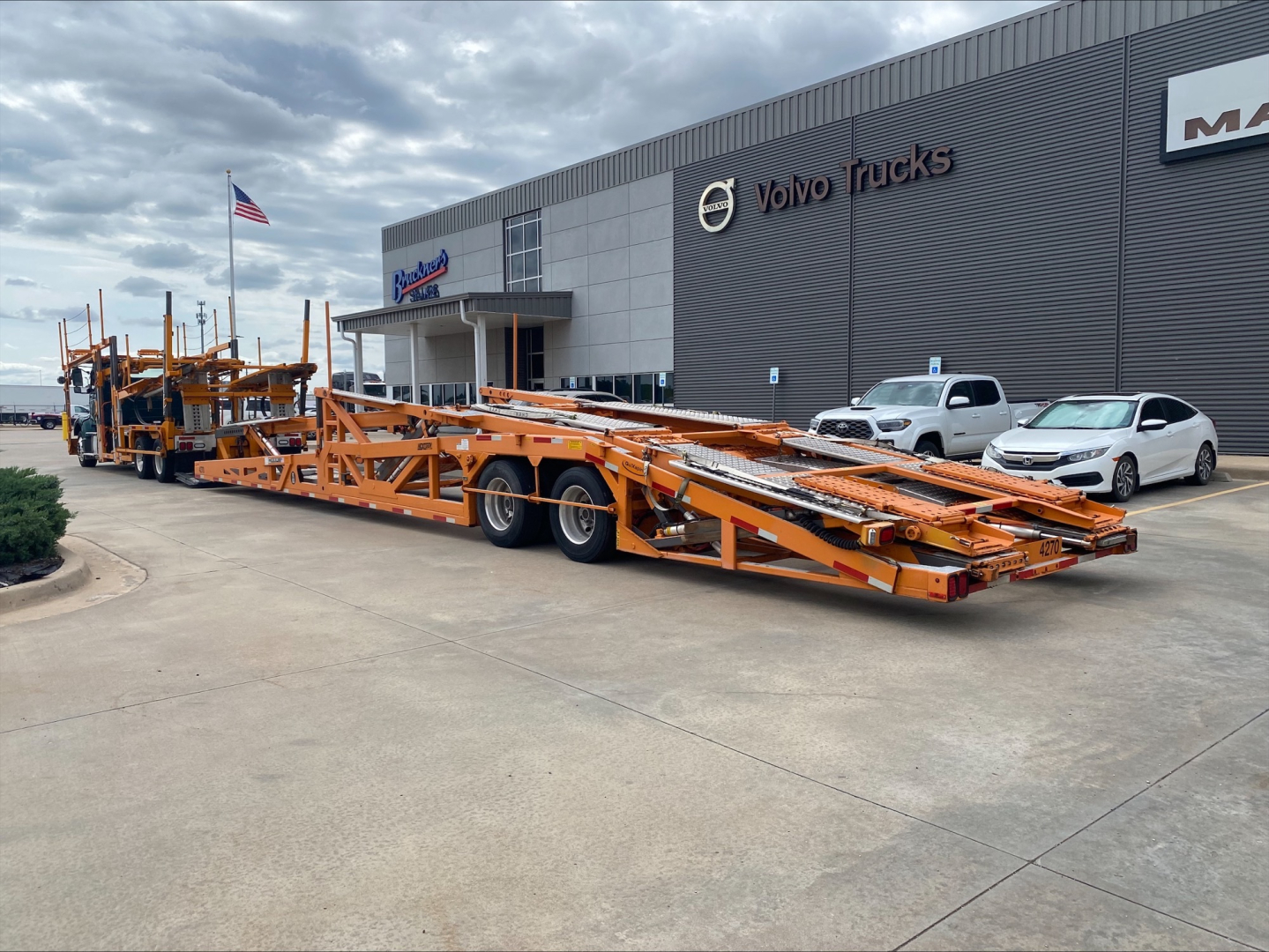 Long orange car hauler trailer attached to a Volvo VAH auto carrier in front of a Volvo Trucks dealership.