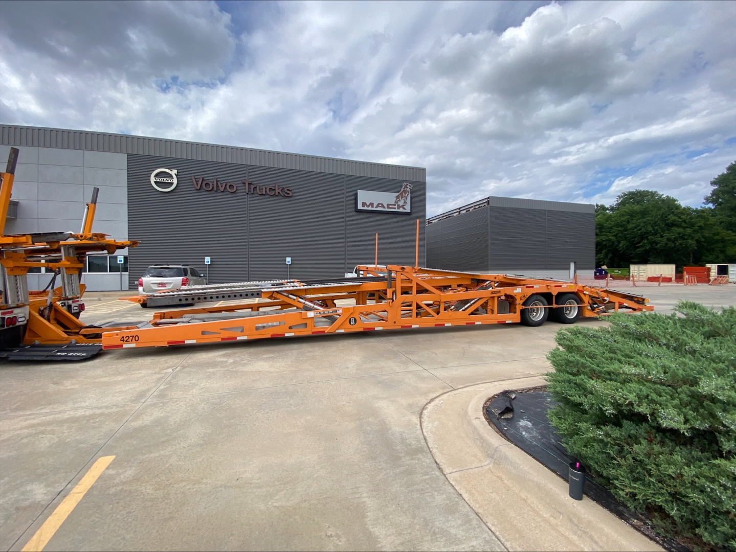 Truck carrier trailer in front of Volvo Trucks building with orange car hauler and stacked equipment, VIN sticker and stock numbers not visible.
