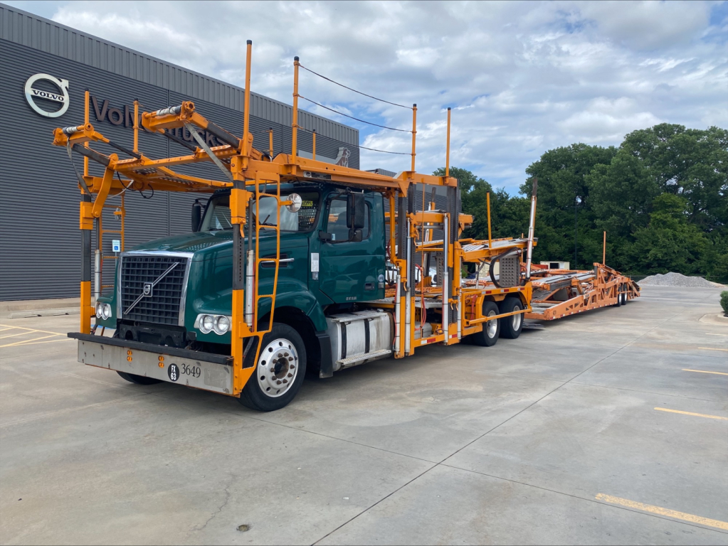 Volvo VAH heavy-haul car carrier truck with orange car deck frame and teal cab, attached to a matching green tractor, long orange loading ramps and equipment, parked outside a dealership.