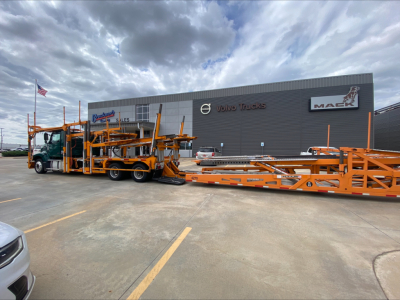 Truck carrier with orange multi-axle trailer loaded with vehicle transport equipment in front of Volvo Trucks/Mack dealership building.