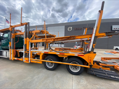Volvo VAH auto carrier truck with two-level car hauler trailer in yellow-orange, side view showing ramps, hydraulic lines, and stacked wheel cradle components.