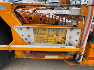 Orange Volvo VAH truck component with metal plate displaying caution label and numbers 9876 54121, hydraulic lines and control levers.