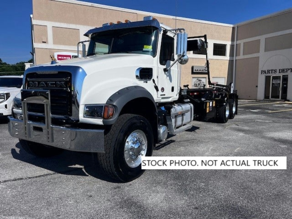 Truck shows Mack GR64F chassis with flatbed in daytime. Front grille, heavy-duty bumper, dual rear axles, chrome fuel tanks, and side mirrors visible.