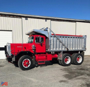 Dump truck with red cab and silver dump bed, three-axle configuration, front grille guard, and raised hoist in a yard.