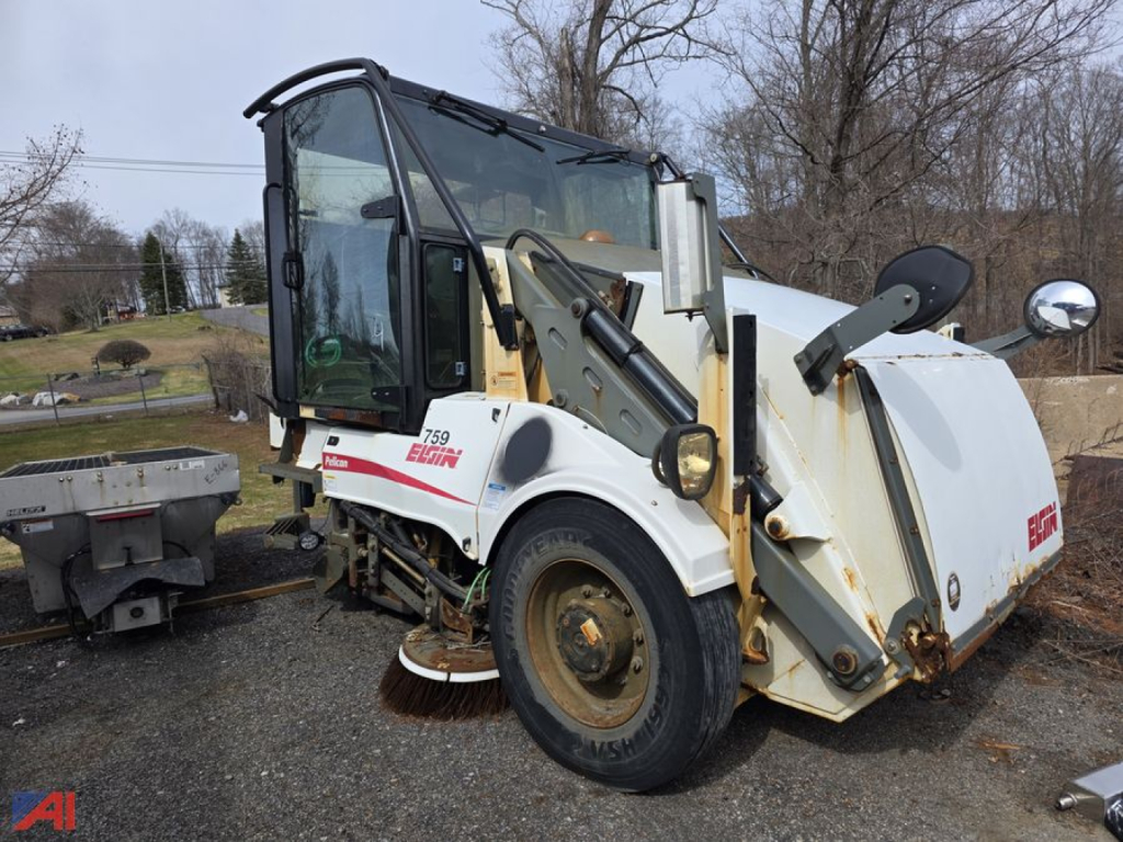 Elgin sweeper truck side view showing side-mounted sweeping arm and brush housing, front cab with windows, white body with red Elgin logo, rusted wheels.