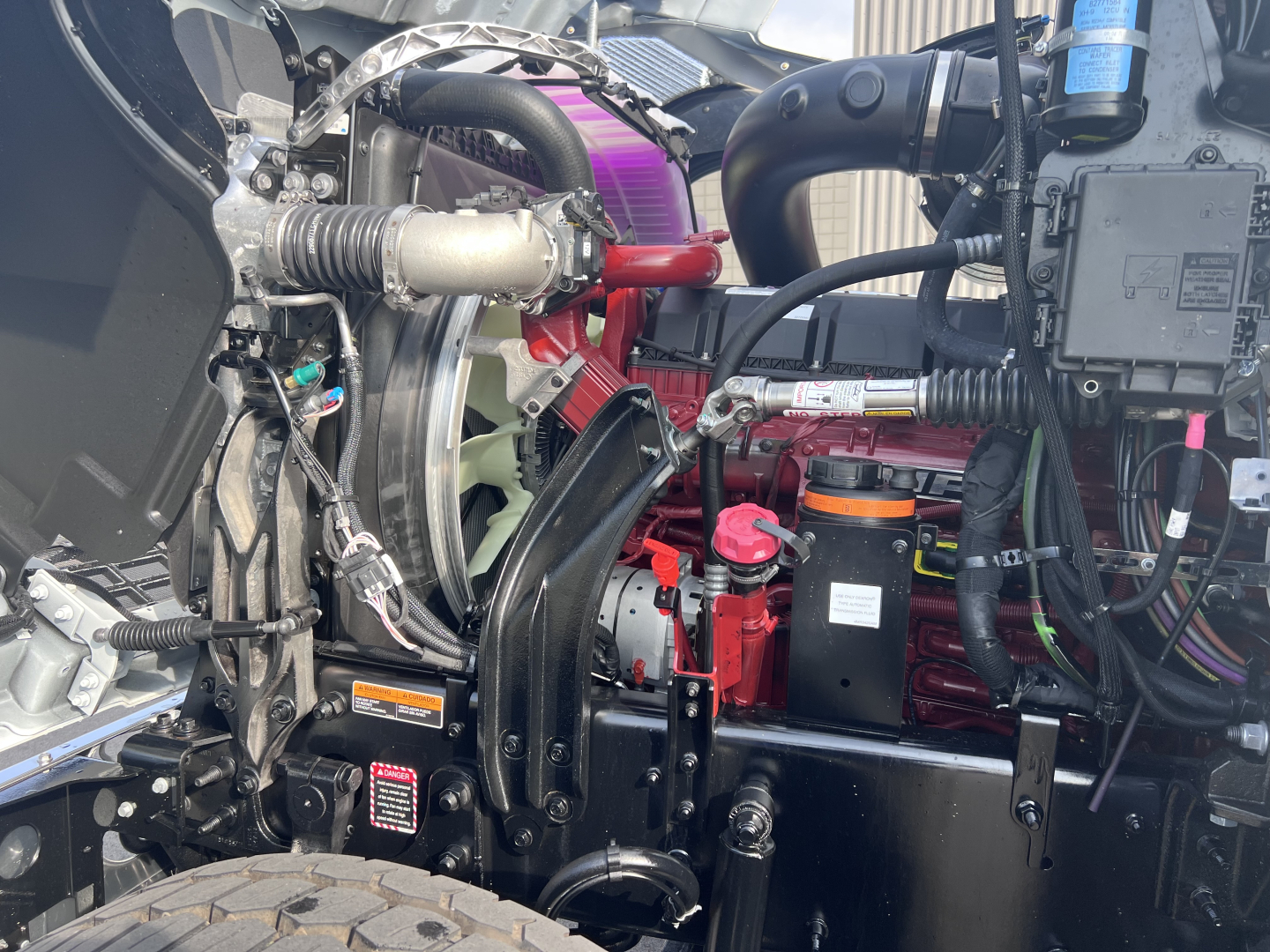 Mack PI64T engine bay showing various components including red engine block, black radiator support, hoses, wiring, and a pink/purple air intake component.